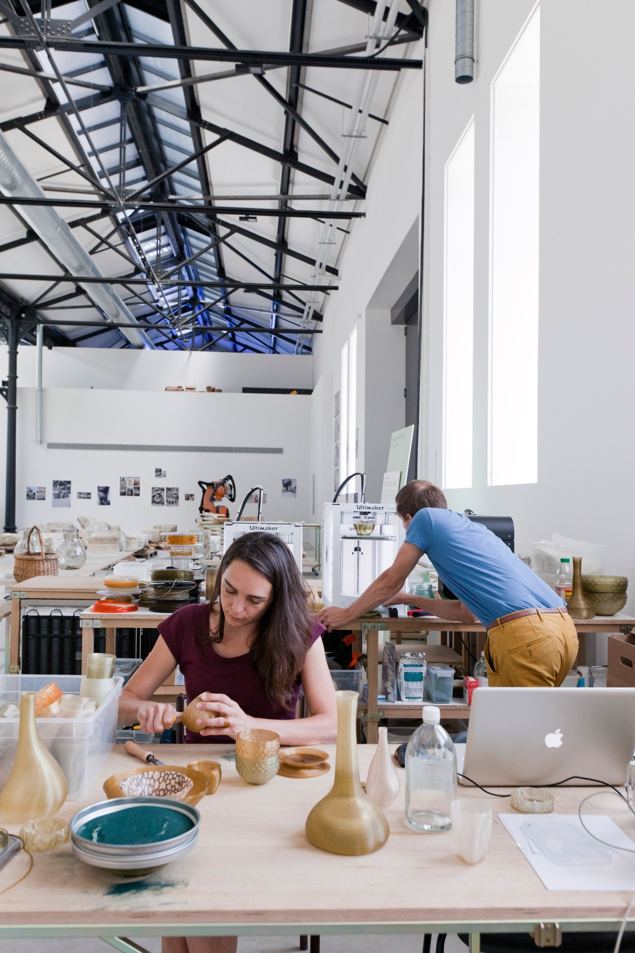 Woman in burgundy shirt working with materials at table while man in blue shirt works at station with 3D printers in warehouse