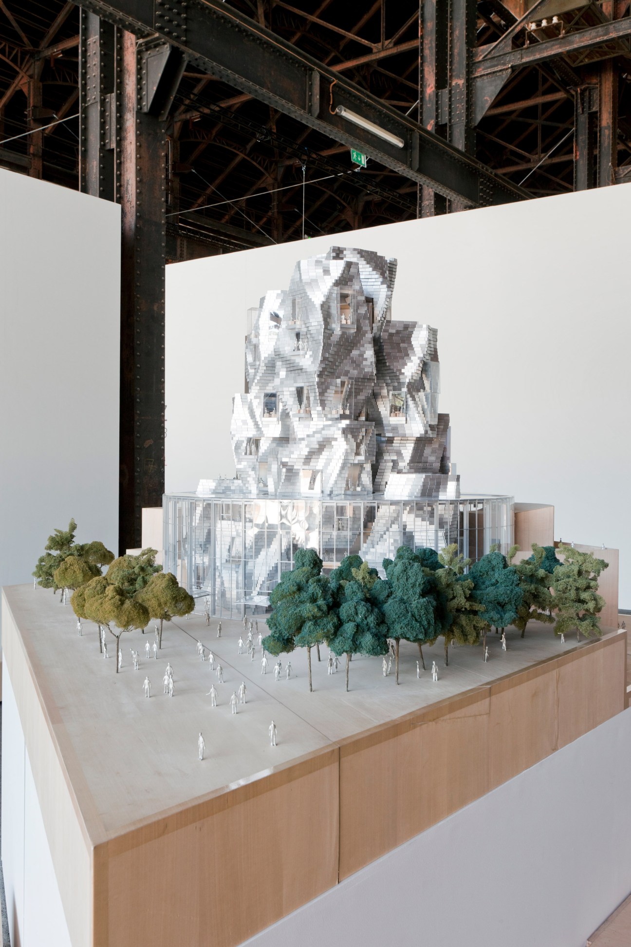 Architectural model of Frank Gehry building with metallic wavy forms surrounded by green trees on display table