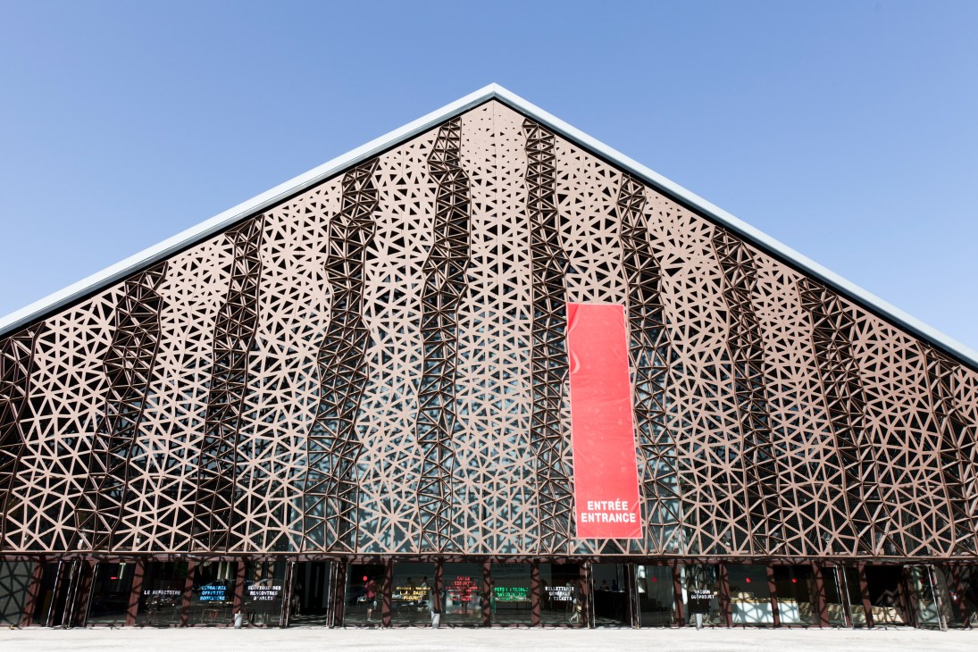 Triangular building facade with geometric perforated metal screening and red entrance sign