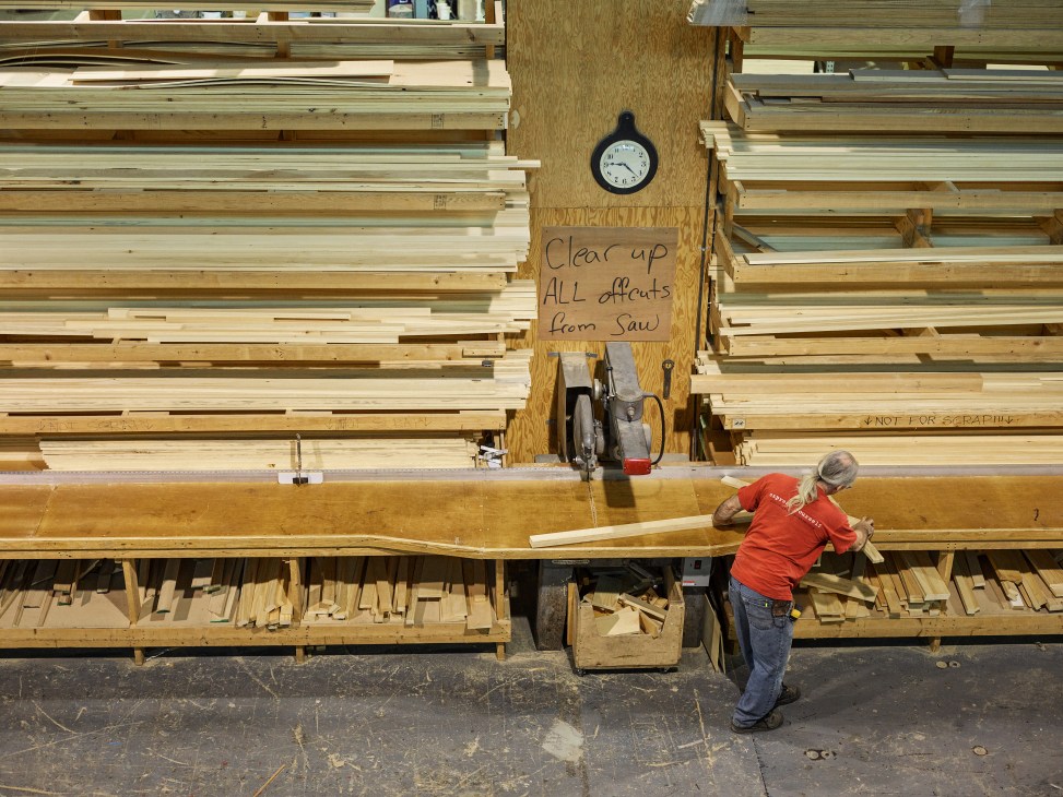 Worker in red shirt organizing wooden lumber and materials stacked against workshop wall with clock and handwritten sign.