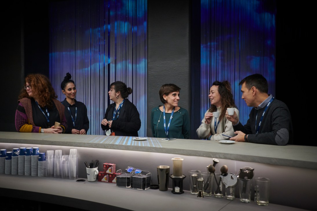 Six people with lanyards socializing around a modern coffee bar with blue lighting and stacked cups