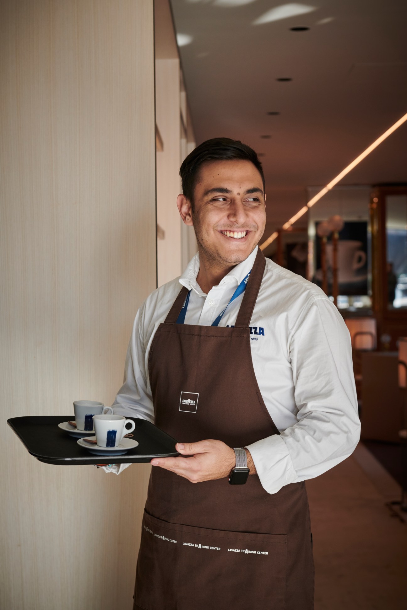 Barman Fabio Andrana smiling while holding a tray with espresso cups in a modern café setting