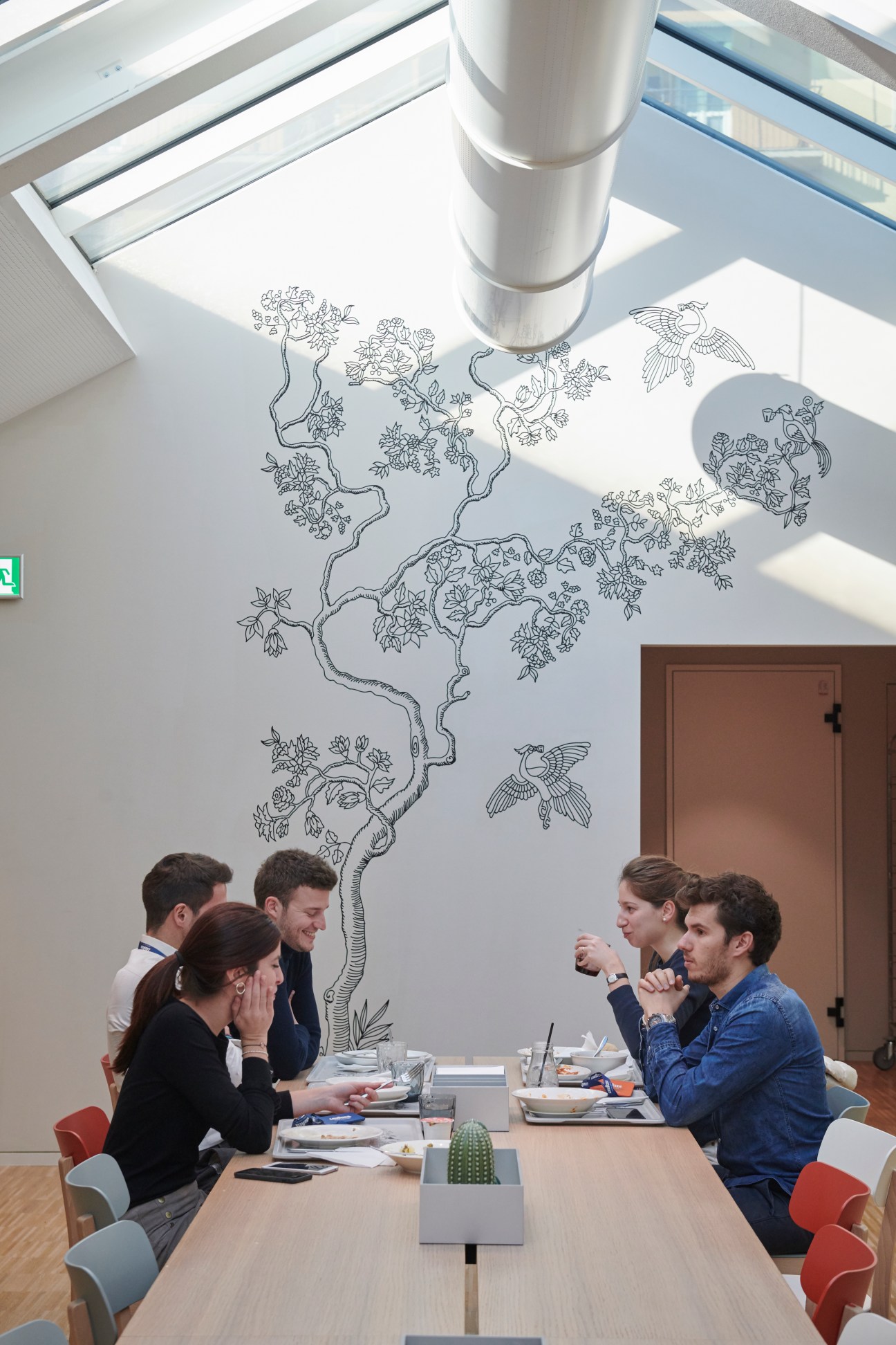 Four people having lunch at a modern table with decorative tree mural on white wall in corporate cafeteria