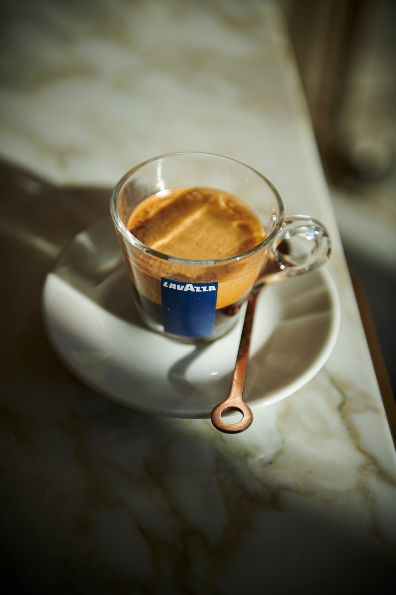 Glass coffee cup with Lavazza branding containing espresso on white saucer with small wooden spoon