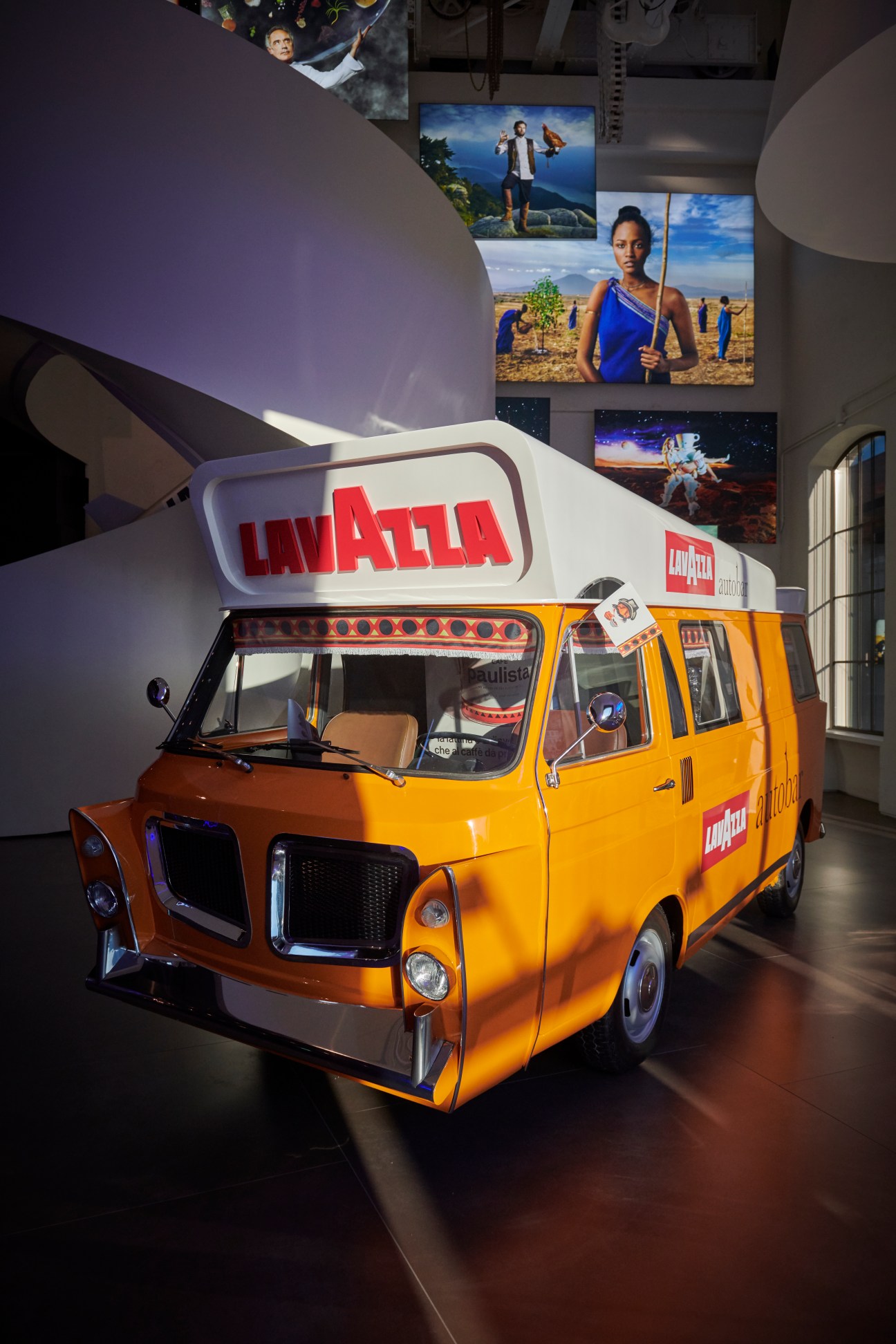 Orange Lavazza coffee truck on display in museum with curved white ceiling and wall screens