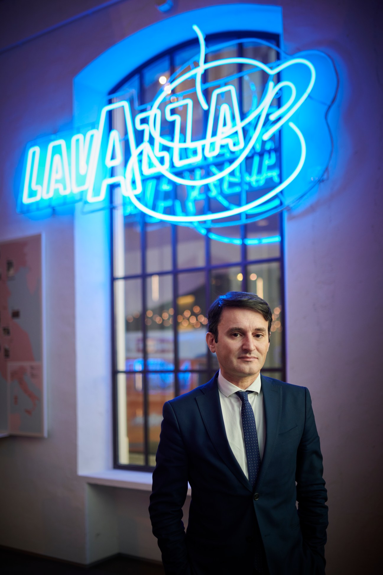 Marco Amato stands in a business suit in front of a large blue neon Lavazza sign