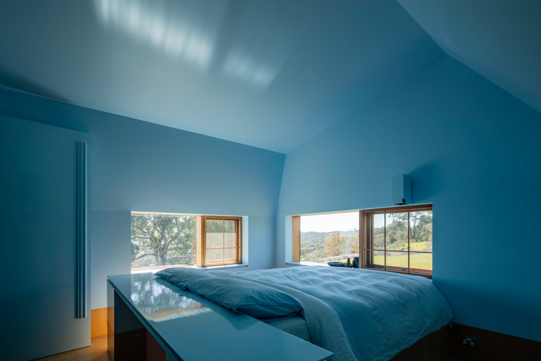 Minimalist bedroom with blue bedding and wood-framed windows overlooking green countryside landscape