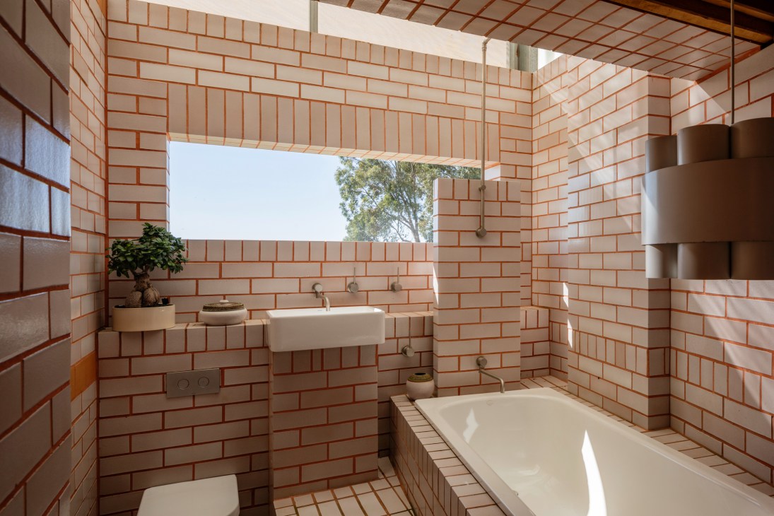 Modern bathroom with cream brick walls, white bathtub, floating sink, and window opening to green trees outside