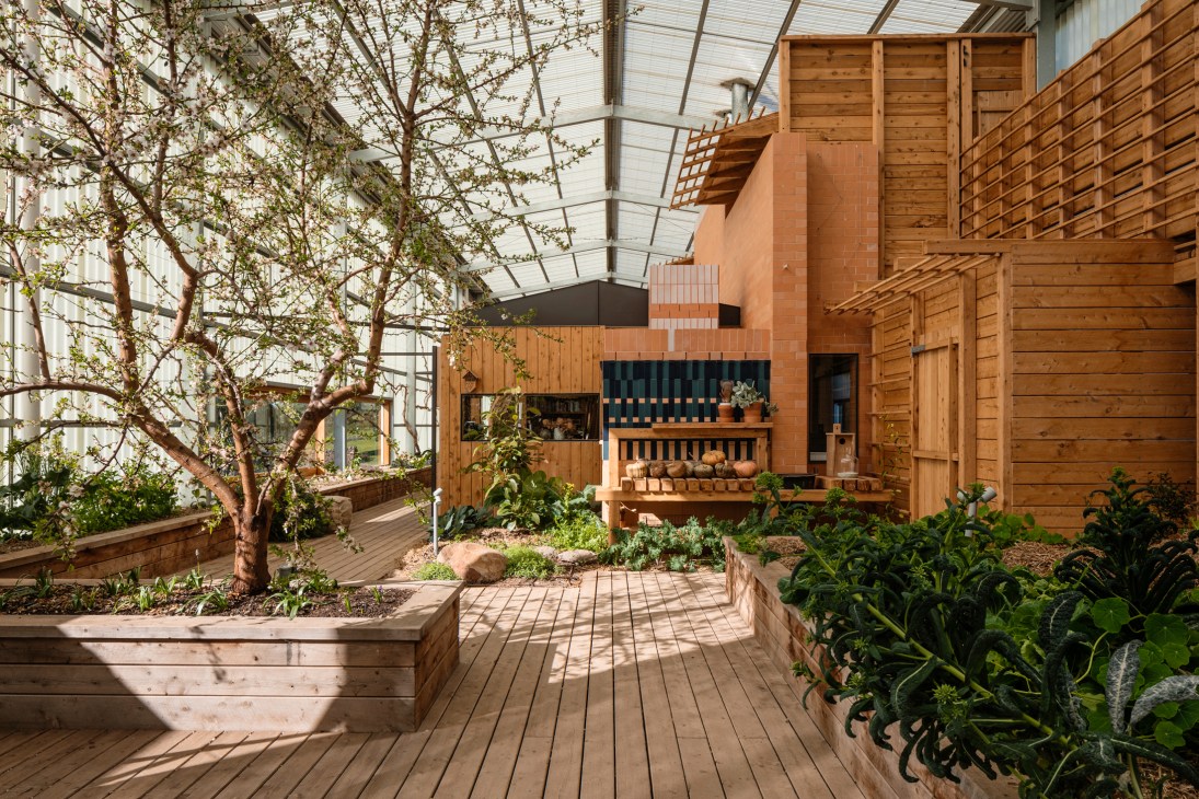 Interior greenhouse space with wooden architecture, raised garden beds with leafy vegetables, and a blossoming tree