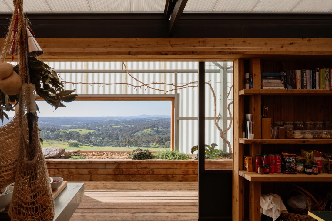 Interior view of wooden house with large window opening to countryside landscape, featuring built-in bookshelves and natural materials