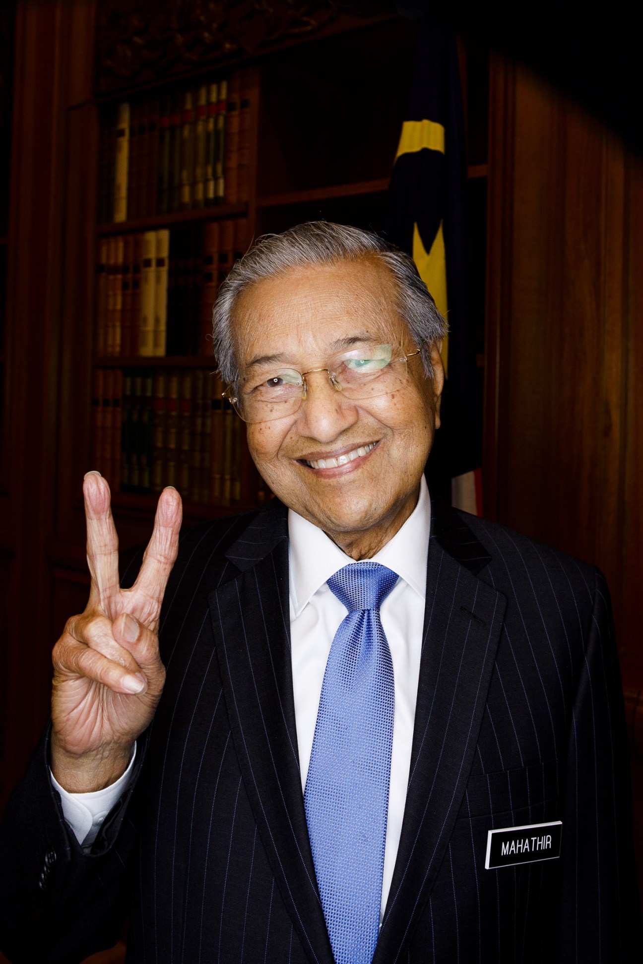 Mahathir Mohamad in a dark pinstriped suit and blue tie, making a peace sign and smiling at the camera.
