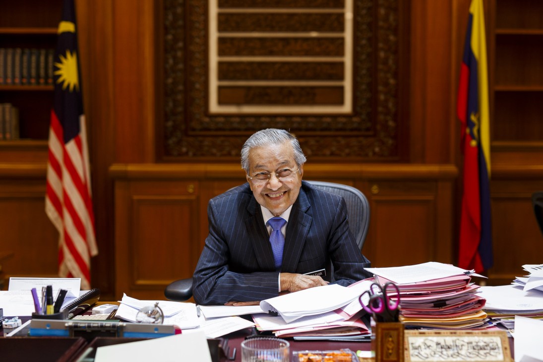 Mahathir Mohamad sits at his desk in his office, surrounded by stacks of documents, with Malaysian flags visible in the background.