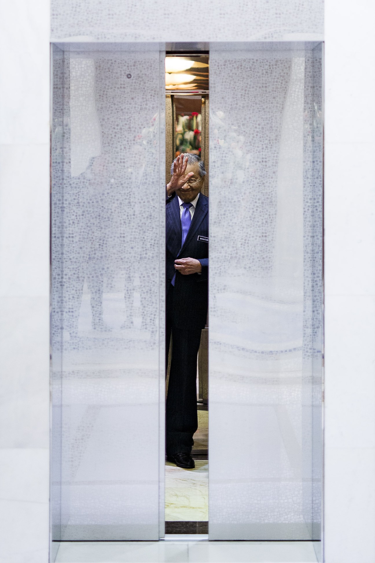 Mahathir Mohamad in a dark suit and blue tie stands in an elevator with his hand raised.