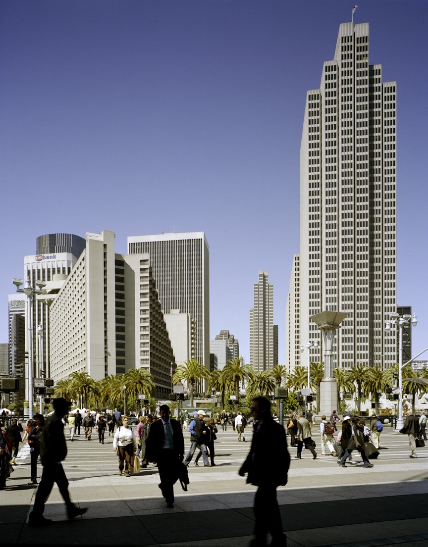 San Francisco's Embarcadero with tall downtown skyscrapers, palm trees, and pedestrians in a public plaza under clear blue sky.
