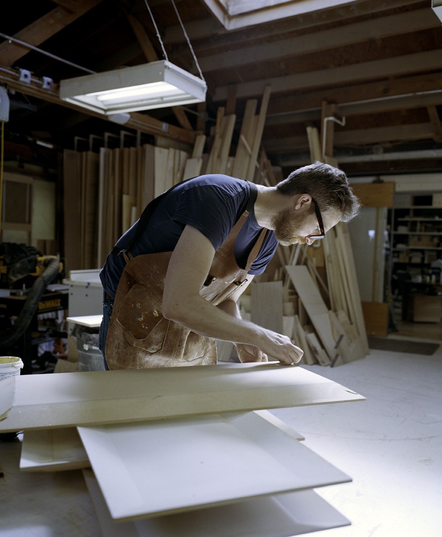 Craftsman working on white furniture pieces in a bright workshop with wooden materials and tools visible in the background.