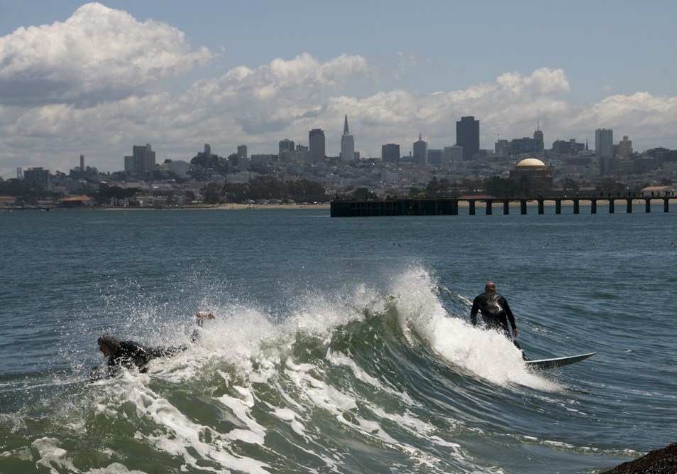 Surfers in wetsuits ride waves off San Francisco's Pacific coast with the city skyline and pier visible in the background.