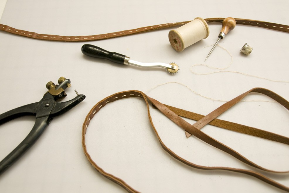 Craft tools and materials including leather straps, thread, needle, punch tool, and metal hardware on white background.