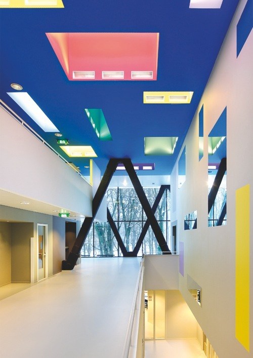 Interior hallway with colorful ceiling lights, white walls, large black X-shaped structural element, and windows overlooking trees.