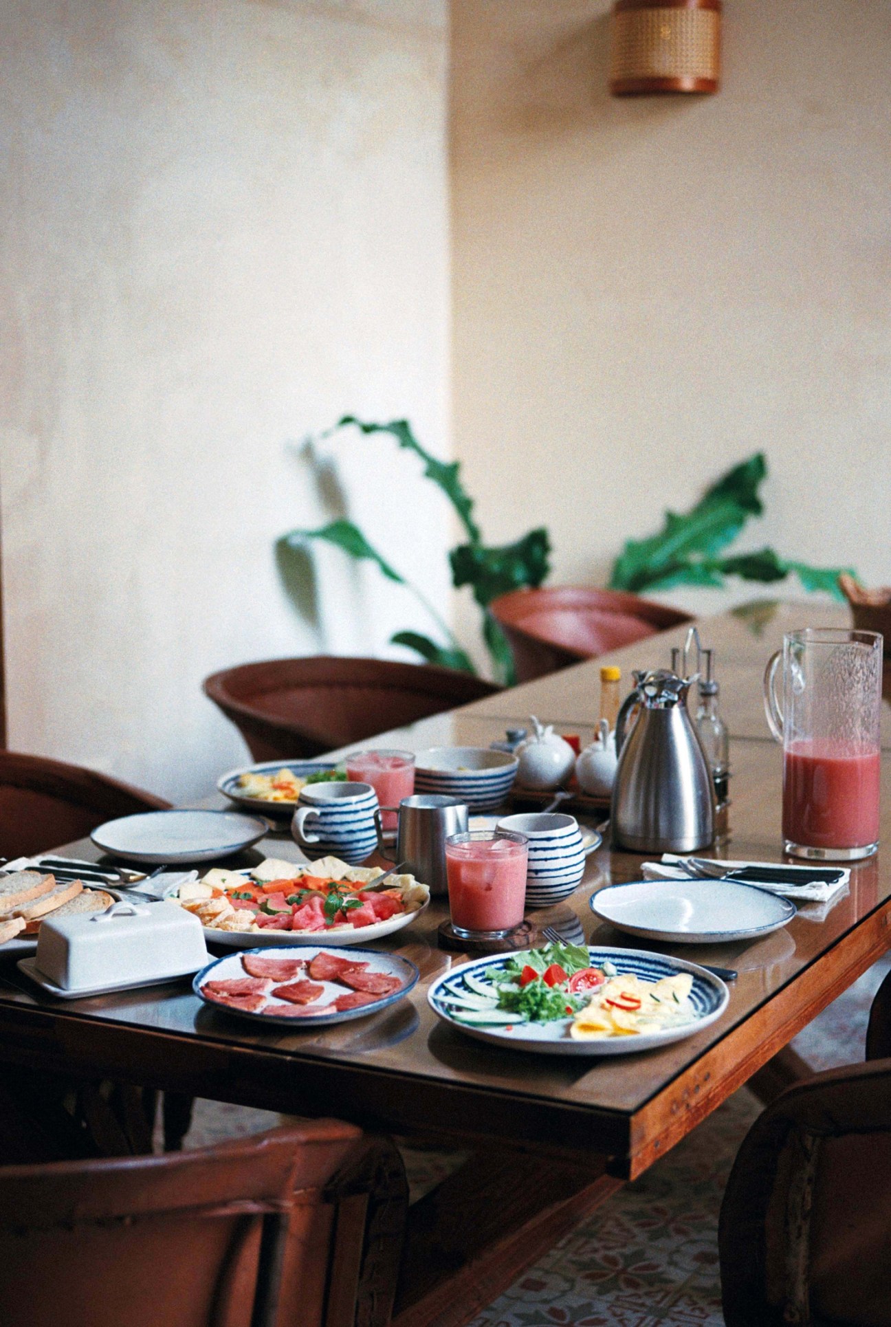 Breakfast table set with plates, glasses of pink juice, coffee pot, and food platters in dining room