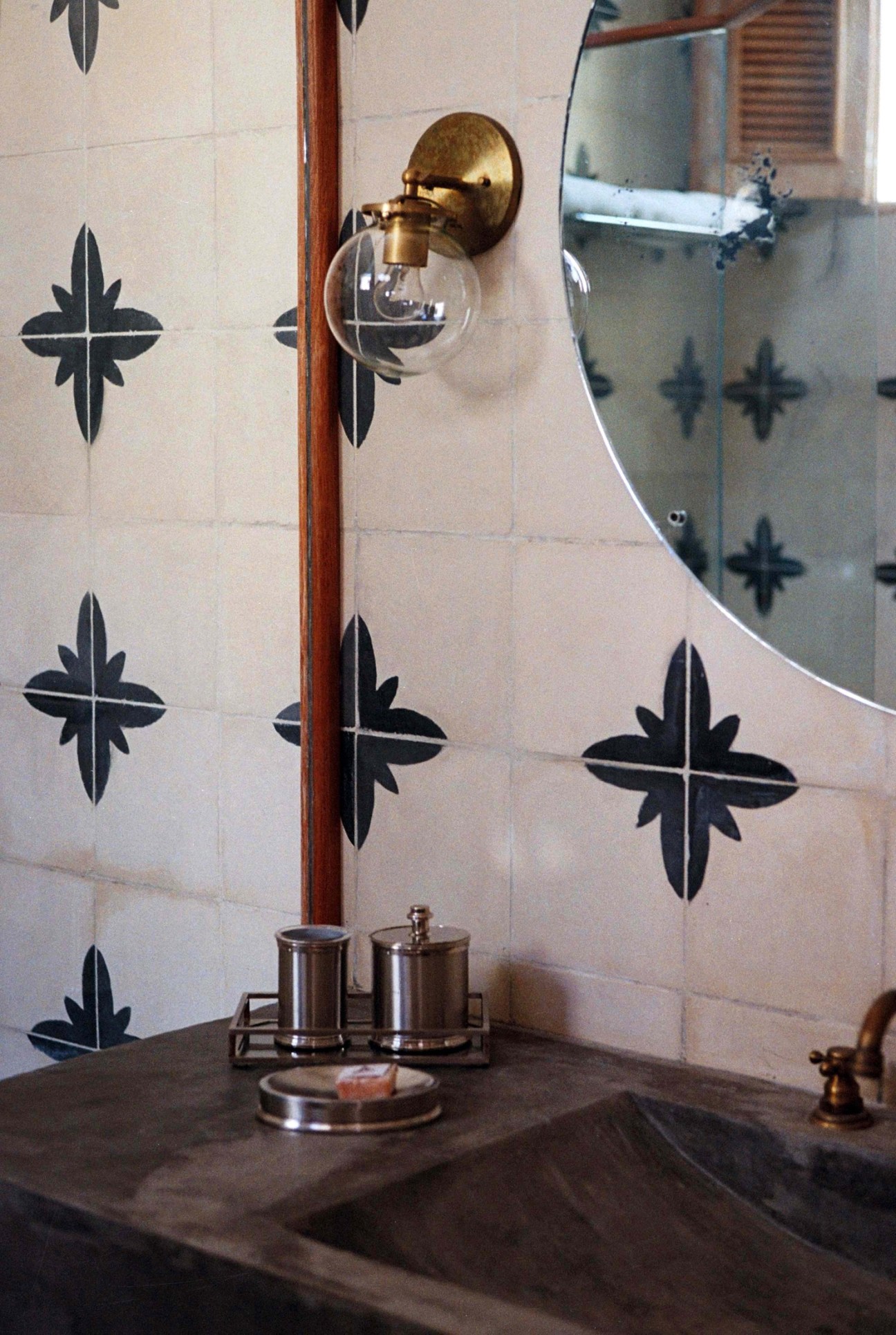 Bathroom vanity with handcrafted patterned tiles, brass fixtures, round mirror, and chrome accessories