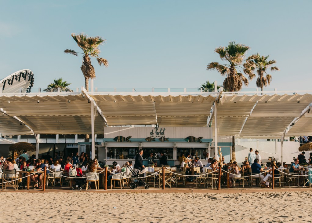 Beachgoers dining at tables under beige canopies at a modern beach restaurant with palm trees and clear sky.