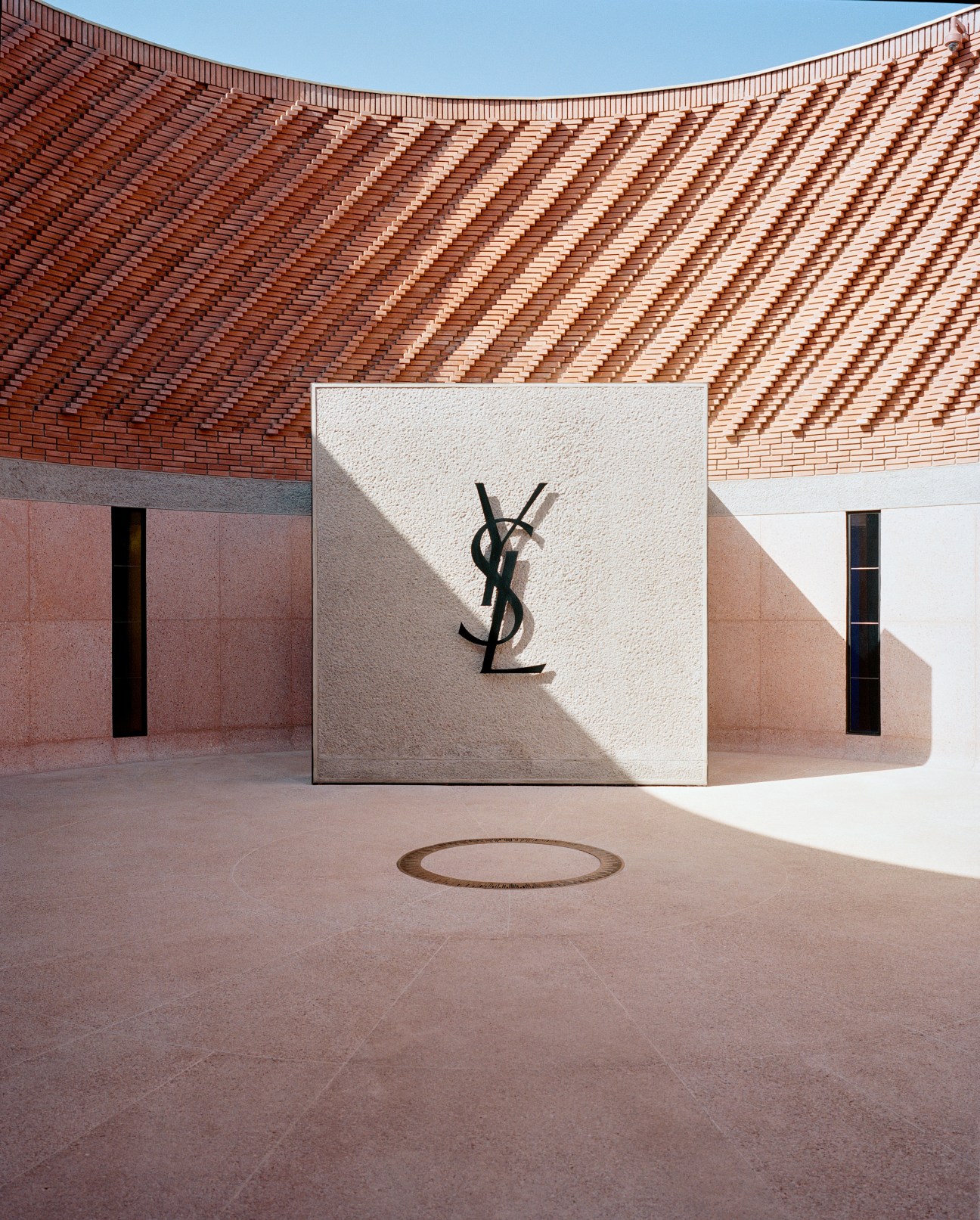 Circular courtyard of Marrakech's Yves Saint Laurent museum with red brick ziggurat architecture and YSL logo on white wall.