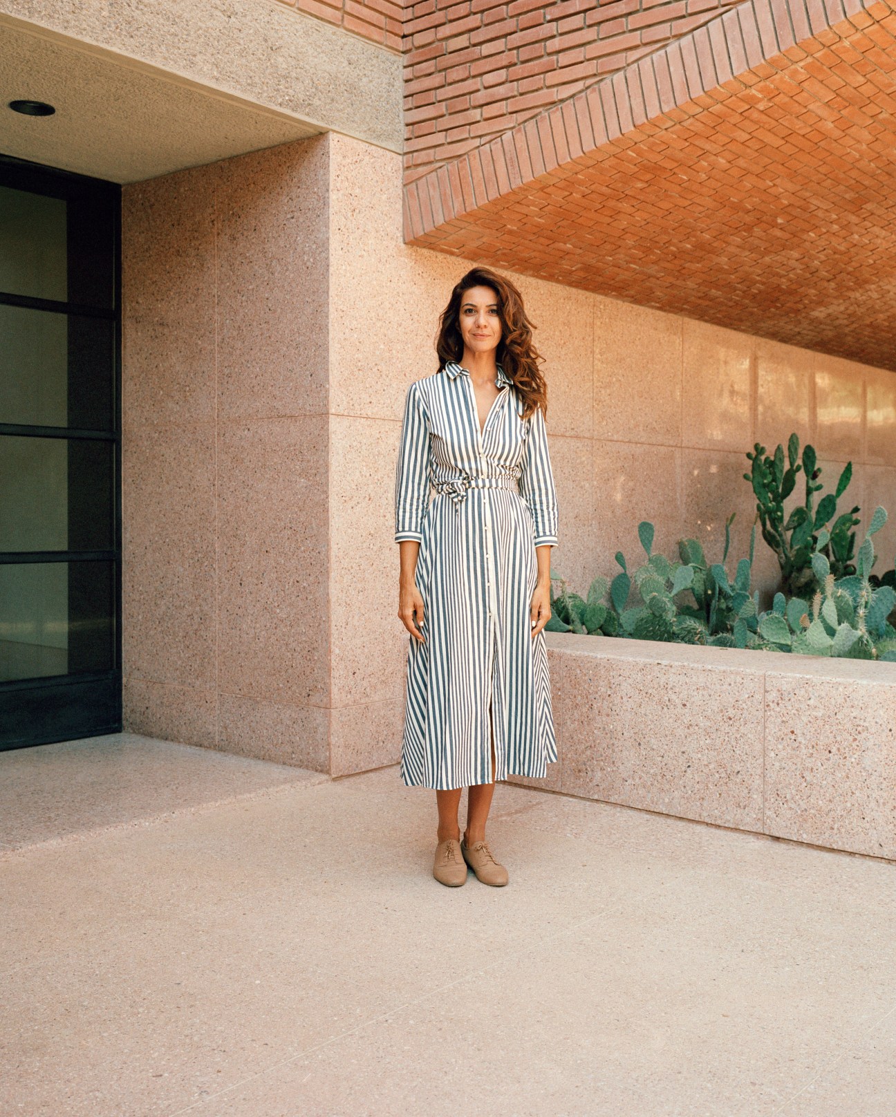 Woman in striped shirt dress and tan boots standing in modernist courtyard with terracotta brick architecture.