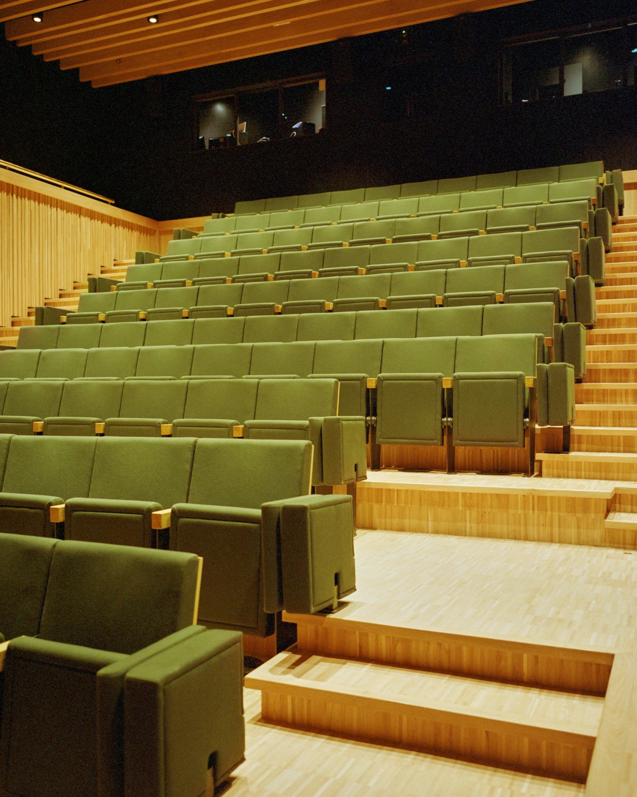 Interior of a modern concert hall with green upholstered seats, wooden steps, and warm wood paneling.
