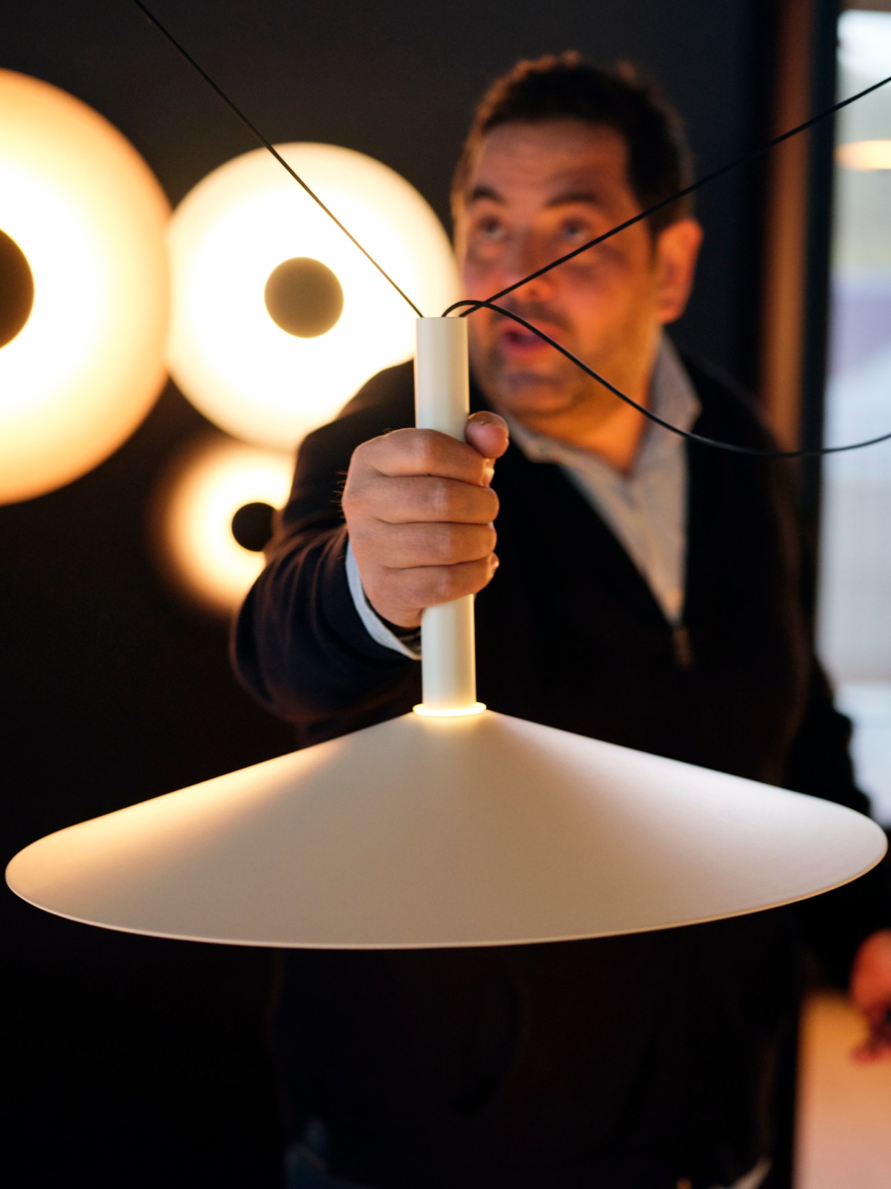Javier adjusts a white pendant lamp with a wide flat shade in front of illuminated circular wall fixtures