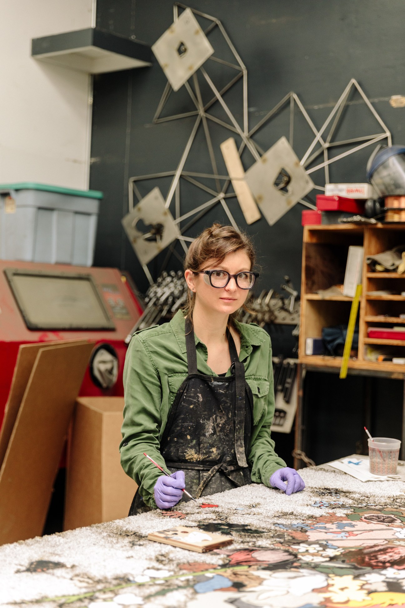 A fabricator in a green jacket and purple gloves works at a table in an art studio.
