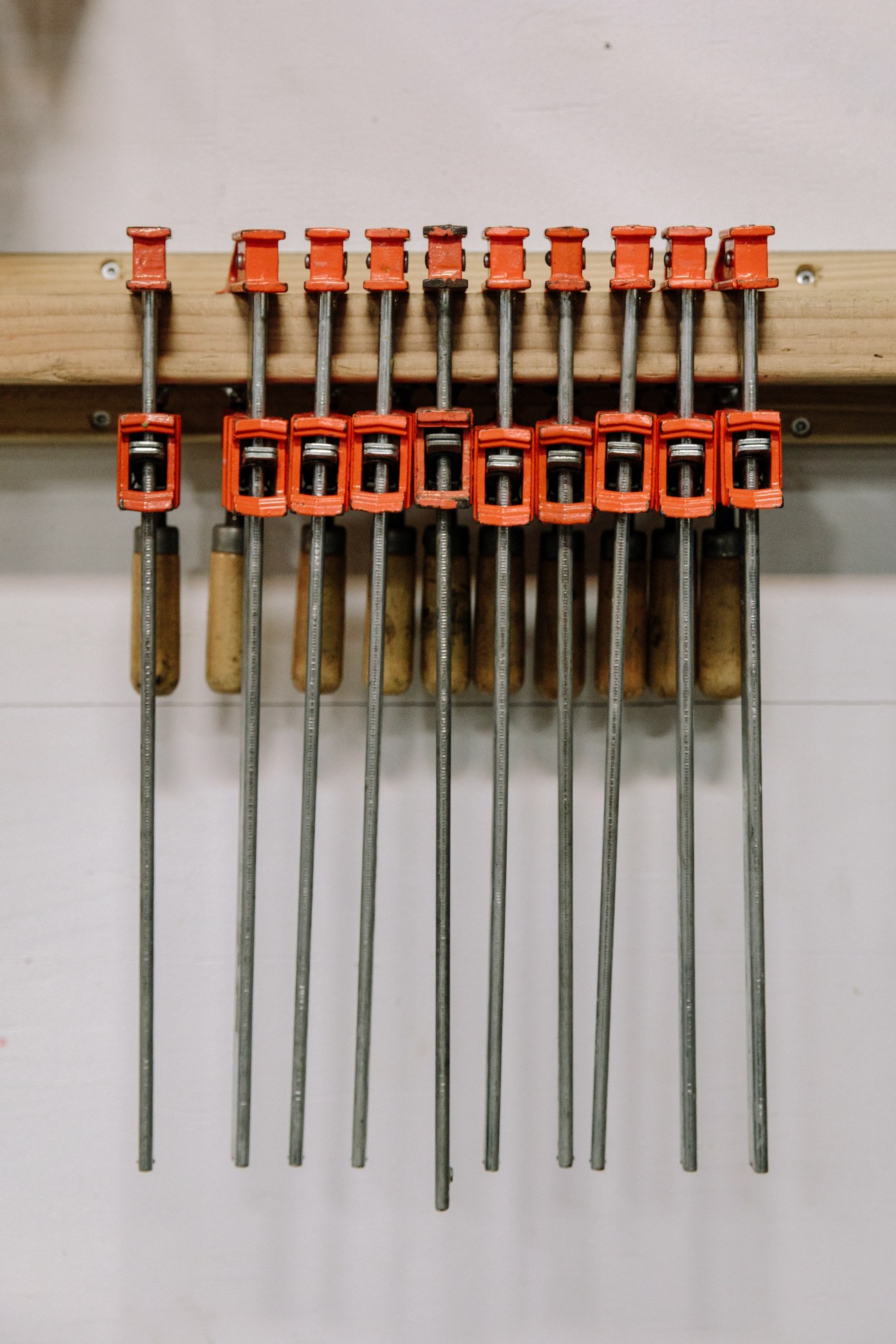 Row of orange metal clamps with wooden and metal handles mounted on a wooden rail in an artist's studio.