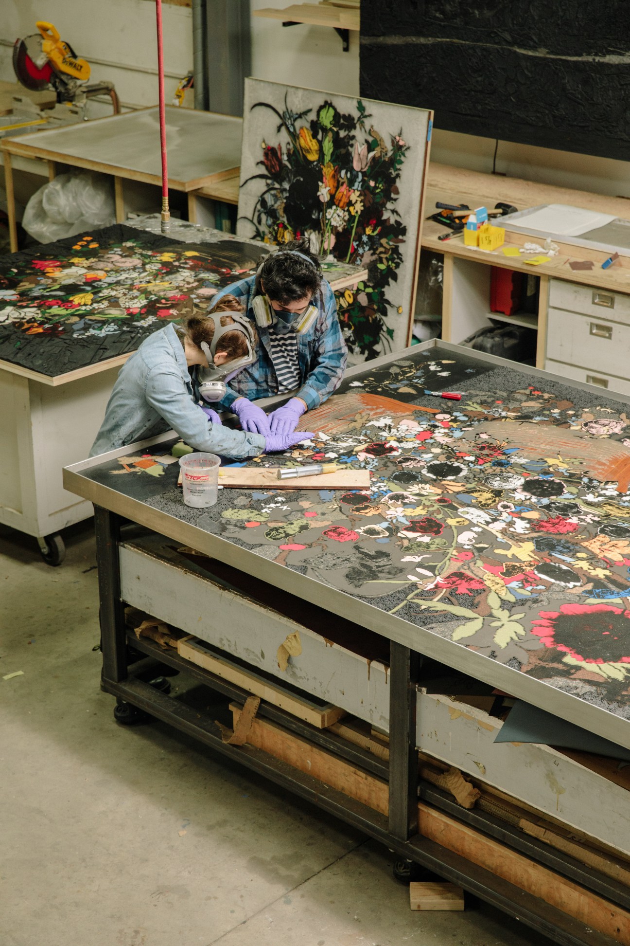 A fabricator wearing protective gear works on colorful floral Formica cutouts at a large studio table.