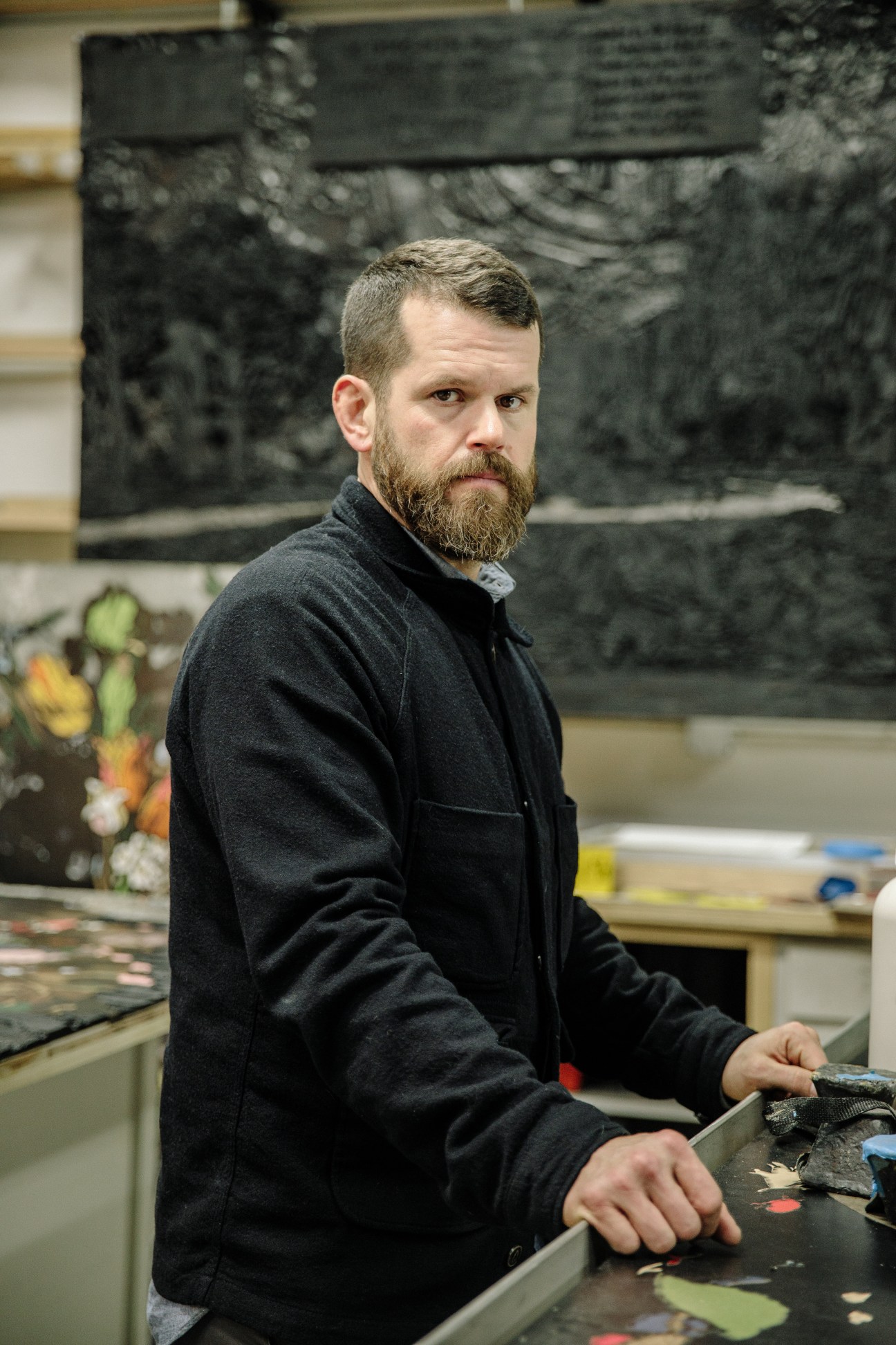 Artist Matthew Day Jackson in his studio, standing at a work table with blackened sculptural works visible behind him.
