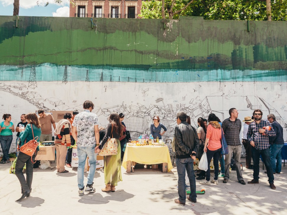 Community gathering at Campo de Cebada with people around food tables and street art mural backdrop.