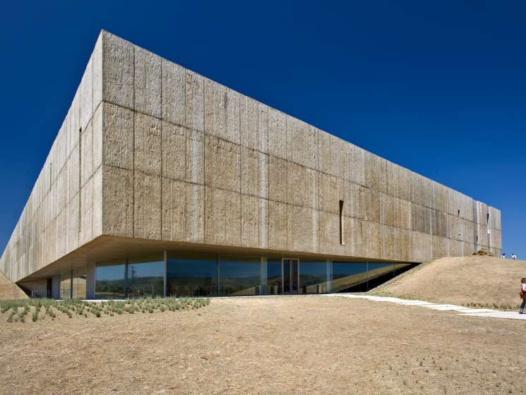 Modern stone museum building with glass windows nestled into a hillside under clear blue sky.