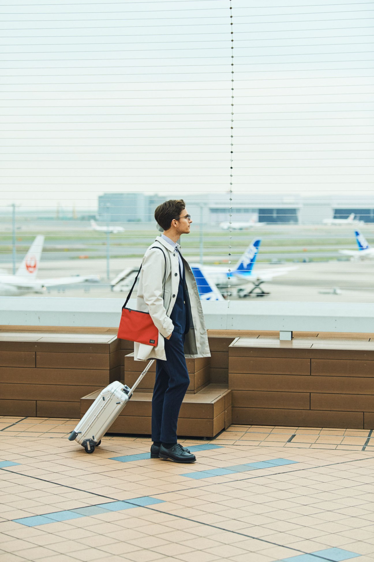 Man in beige coat and navy suit with red shoulder bag standing at airport terminal overlooking runways.