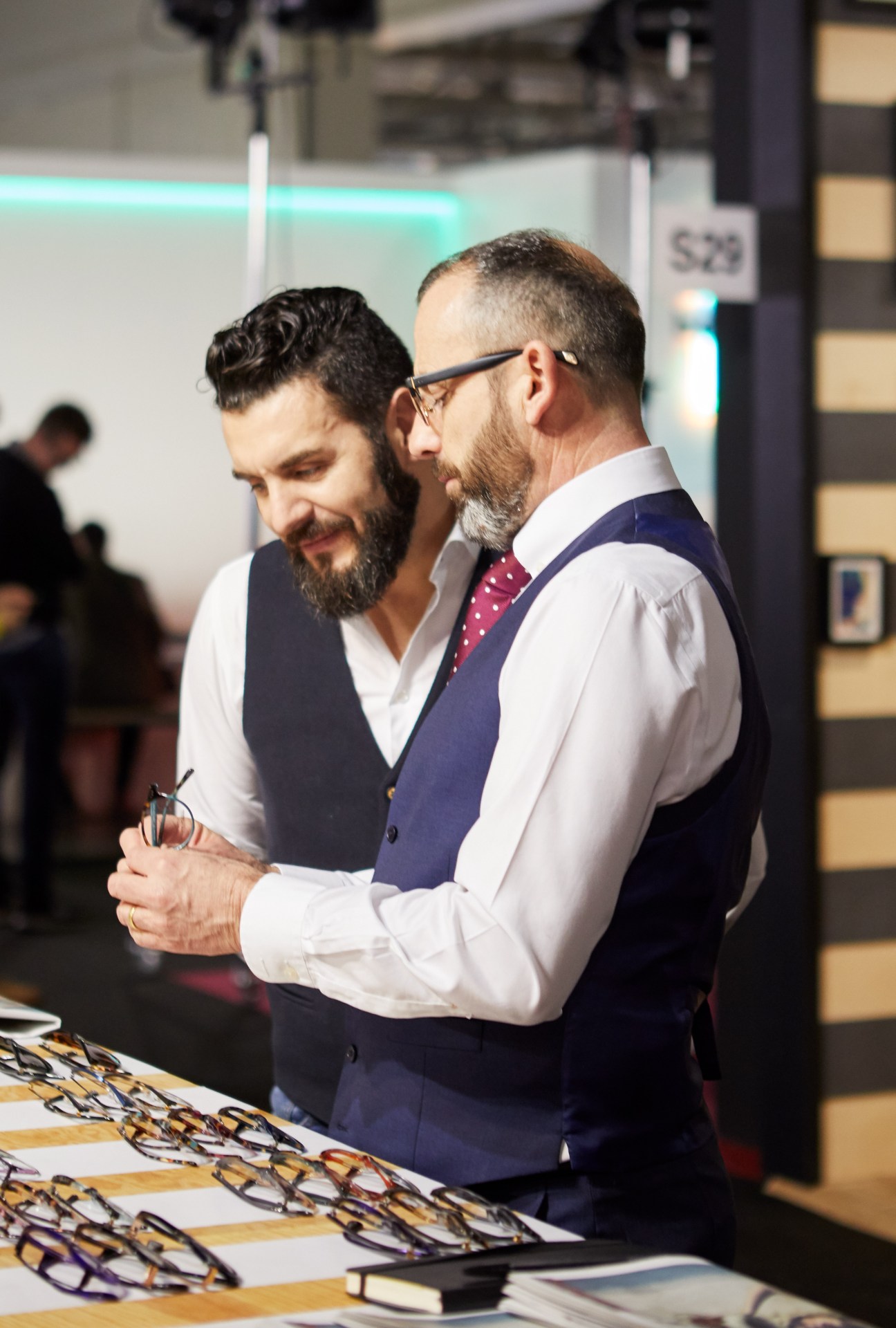 Two men examining eyewear at a trade show booth, one wearing glasses and a navy waistcoat.