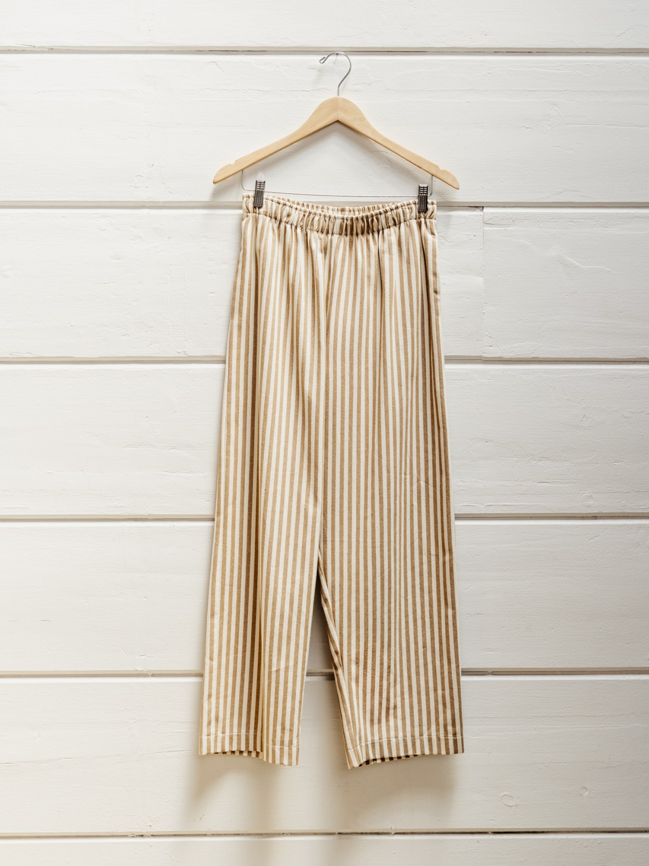 Striped drawstring trousers in tan and cream by Or hung on a wooden hanger against white shiplap wall.
