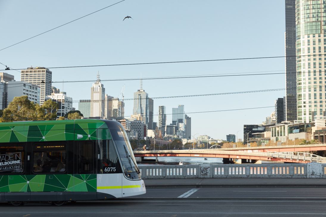 A green and white Melbourne tram crosses the Yarra River with the city skyline in the background.