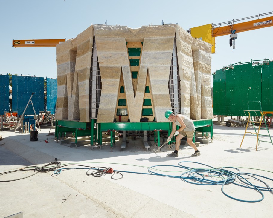 A stone tower section under construction at an outdoor building site in Galera, with a worker and industrial cranes nearby.