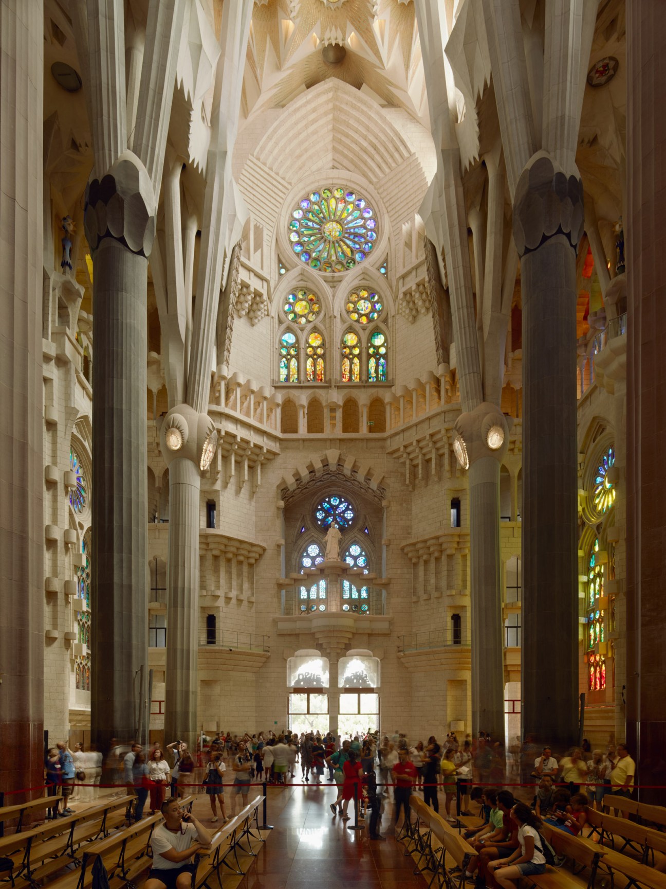 Interior of Sagrada Família basilica with visitors and worshippers during mass, showing soaring columns and stained glass windows.