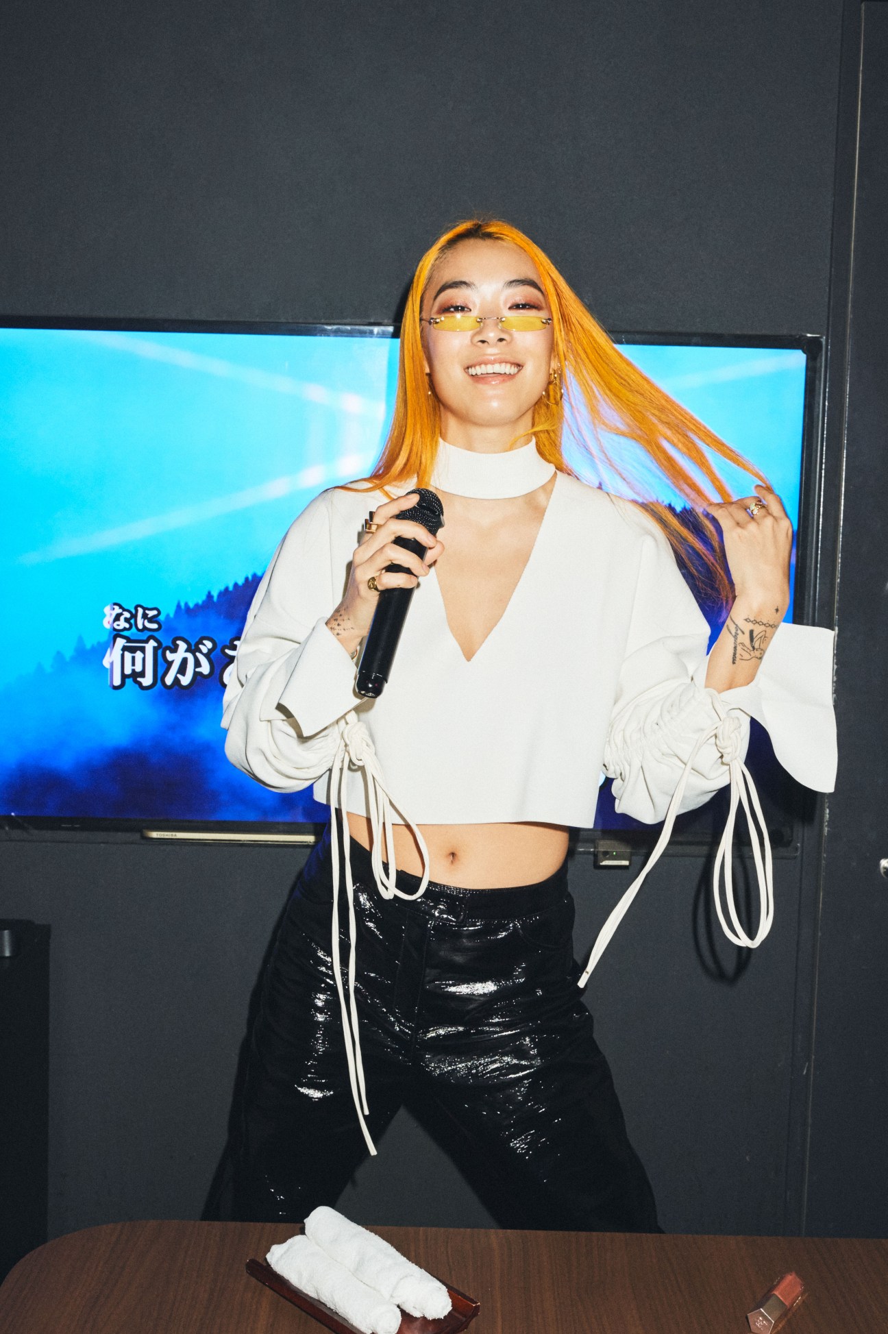 Rina Sawayama performing with a microphone, wearing a white cropped top and black sequined pants.