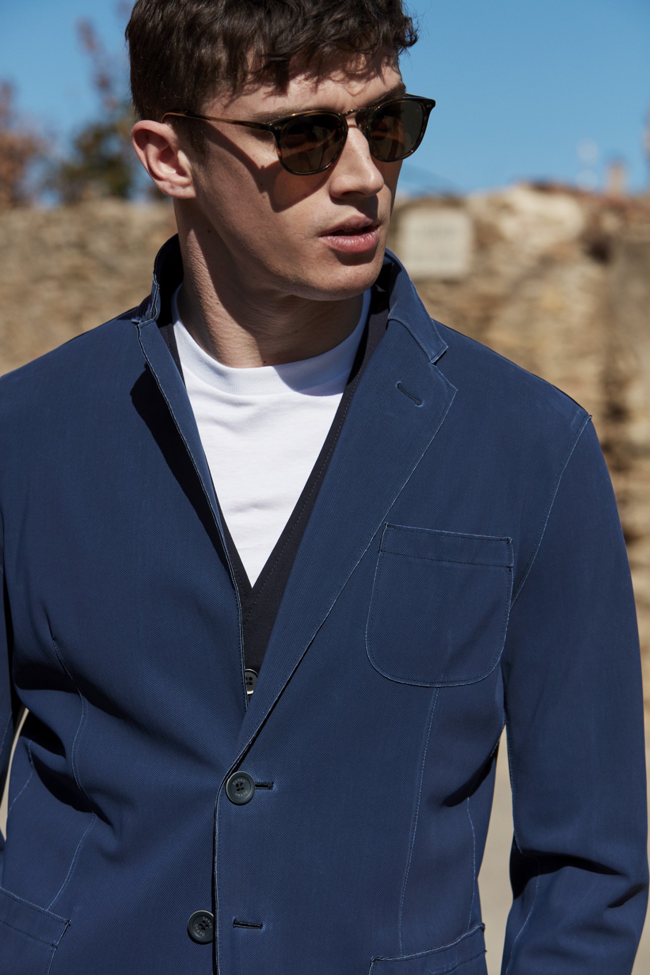 Man wearing navy Herno jacket, Barena Venezia waistcoat, Prada t-shirt and Oliver Peoples sunglasses in desert setting.