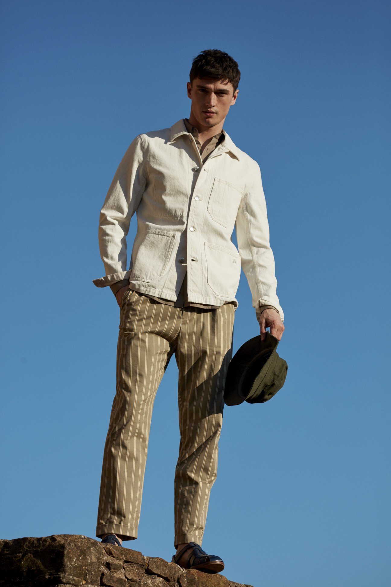 Male model wearing a cream jacket, pinstriped trousers, and holding a hat against a blue sky.