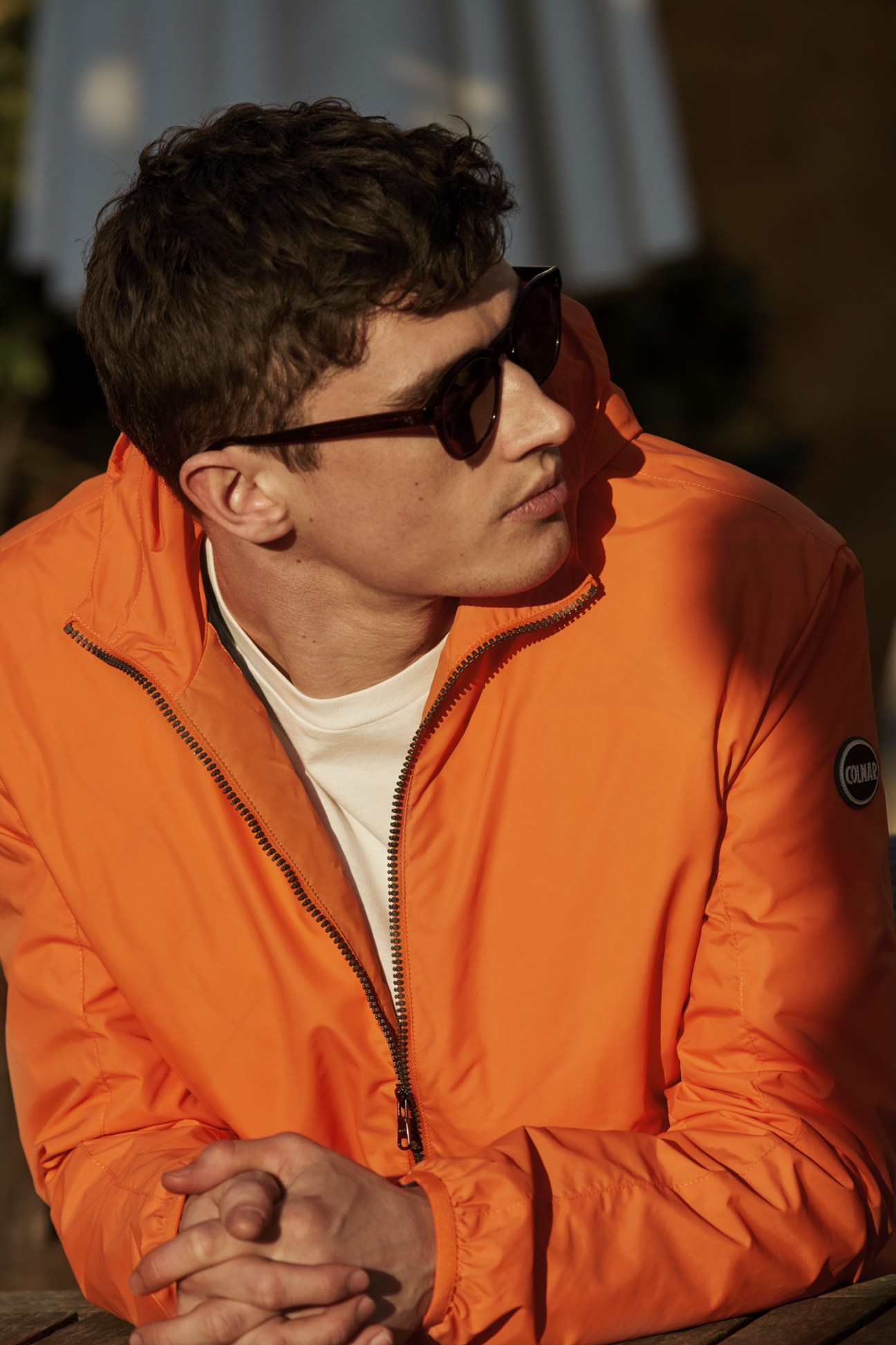 Man wearing bright orange Colmar jacket with Prada t-shirt and Oliver Peoples sunglasses.
