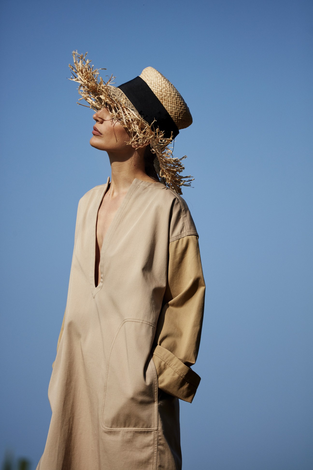 Model wearing a beige tunic by Loewe and a straw hat with black band by Chanel against a blue sky.