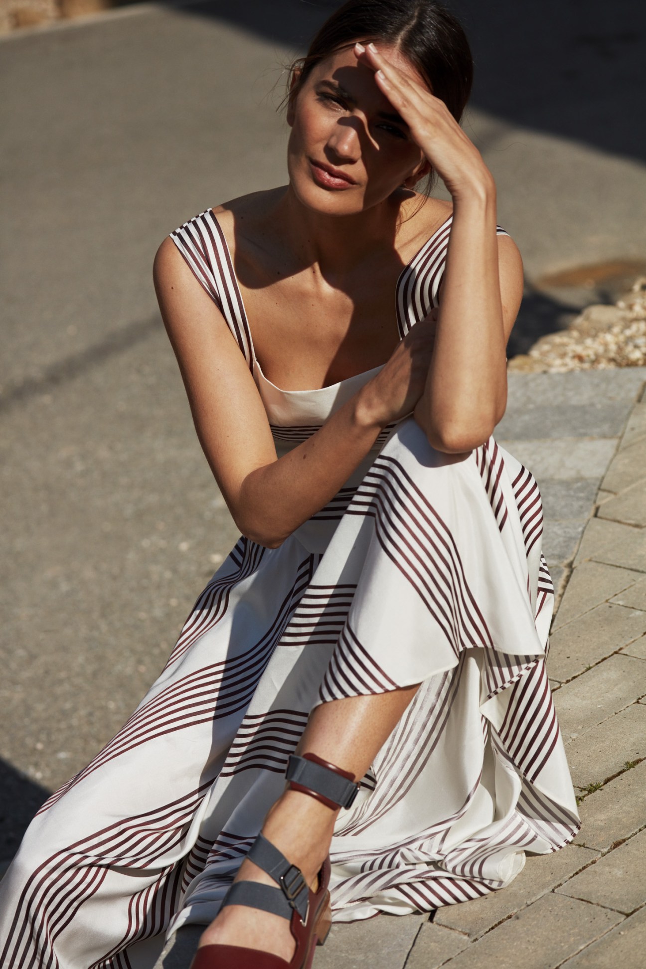 Woman wearing a striped Loro Piana dress and Hermès sandals sitting on pavement in sunlight.