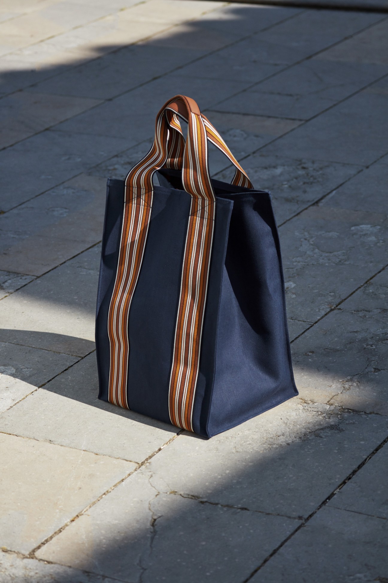 Navy tote bag by Loro Piana with brown and white striped handles on a paved ground.