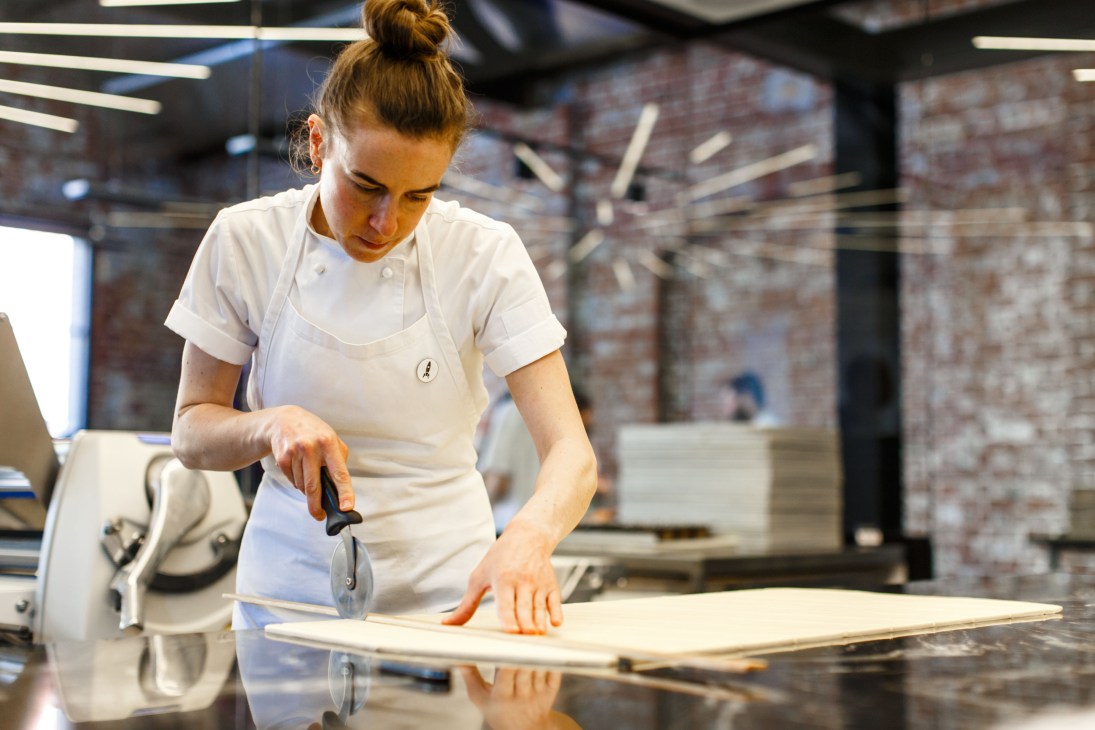 A pastry chef in white chef's coat cutting dough with a pastry wheel in a modern bakery kitchen.