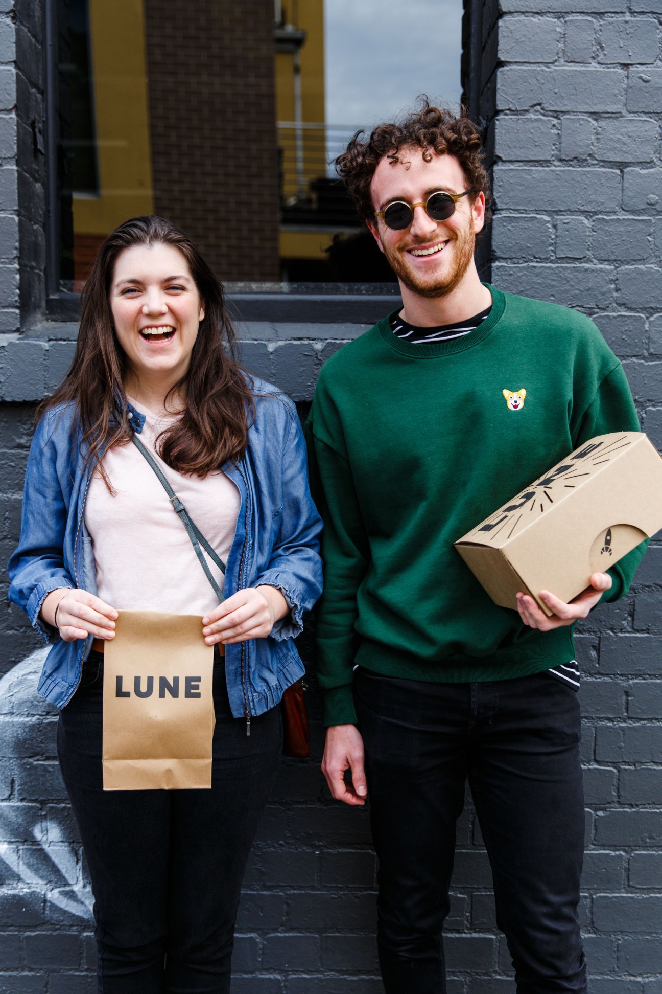 Two young people holding Lune branded paper bags stand smiling against a brick wall.